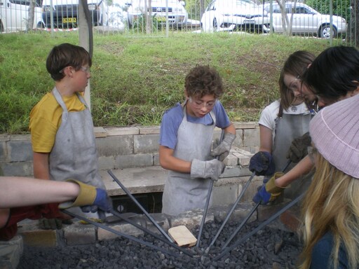 Kindlehill high schoolers collaboratively forge metal at an outdoor hearth, blending practical tool skills with a focus on aesthetics.