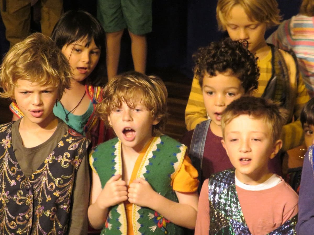 Kindlehill children in colourful costumes sing with soulful focus, performing a theatrical folk story to celebrate community imagination.