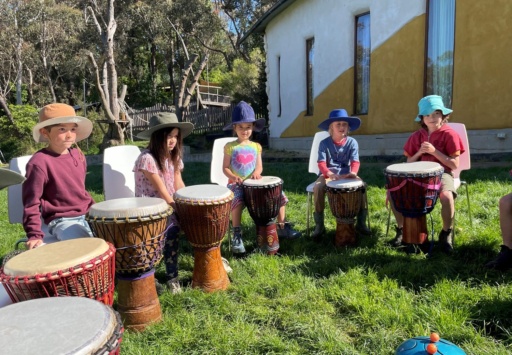 Children practice djembe drumming in a sunlit bushland circle, fostering rhythmic connection and artistic expression within their community.