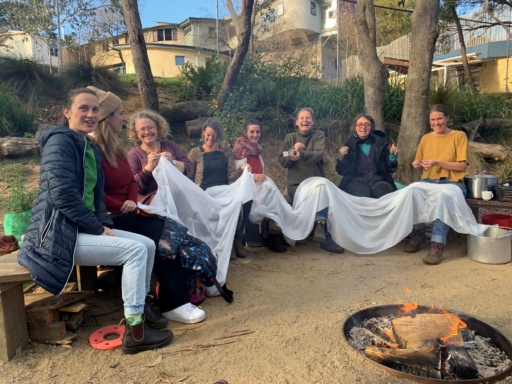 Community members sew together around a bush circle fire, honouring seasonal rhythms and building a village through connection.