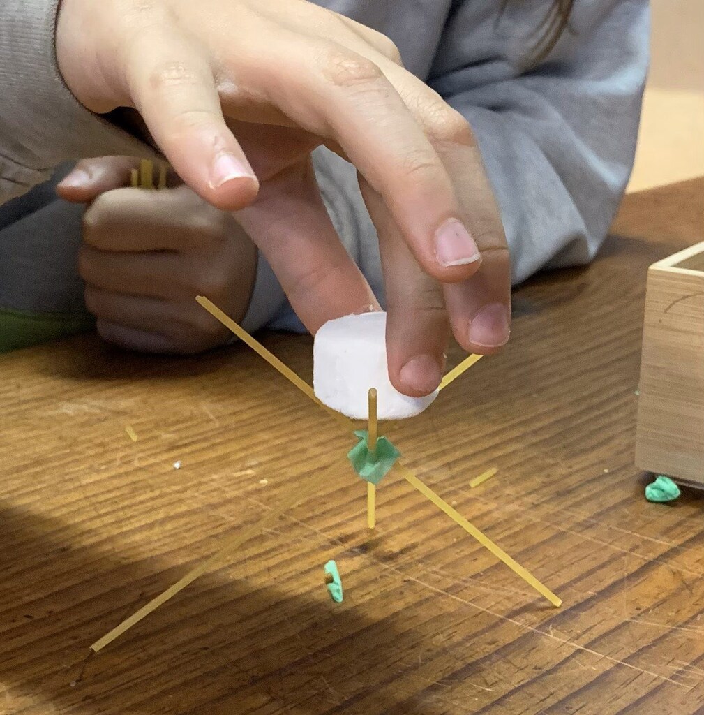 Students practise cooperation and resilience, carefully balancing a marshmallow atop a spaghetti structure during a collaborative STEAM challenge.