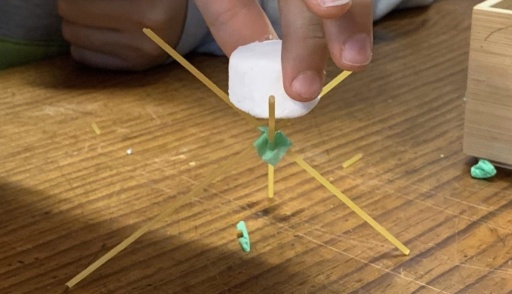 Collaborative hands balance a marshmallow on spaghetti, practising iterative design and creative problem-solving during a Kindlehill STEAM challenge.