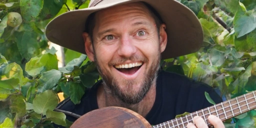 Charlie McGee joyfully shares permaculture tunes amongst green leaves, celebrating the regenerative spirit of the Kindlehill community.