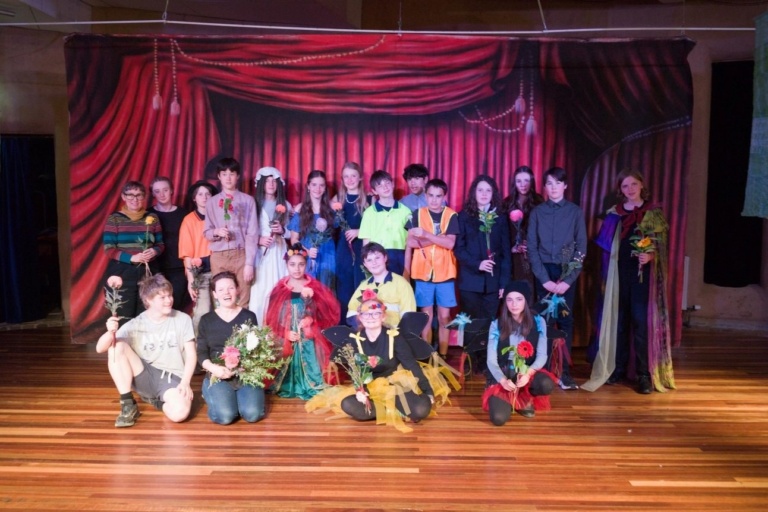 Kindlehill students and teachers celebrate their collaborative Shakespearean theatre performance on stage, holding flowers and showcasing community spirit.