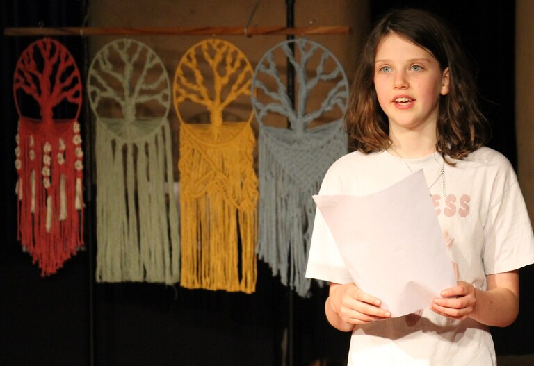 A student confidently presents her Big Project beside vibrant, colourful macrame trees, celebrating her creative passion and growth.