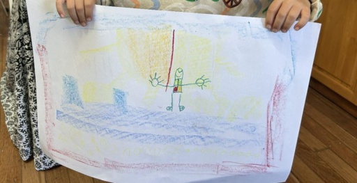 Hands hold a childs colourful crayon drawing of an expressive figure with open arms in a sunlit landscape.