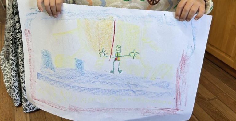 Hands hold a childs colourful crayon drawing of an expressive figure with open arms in a sunlit landscape.