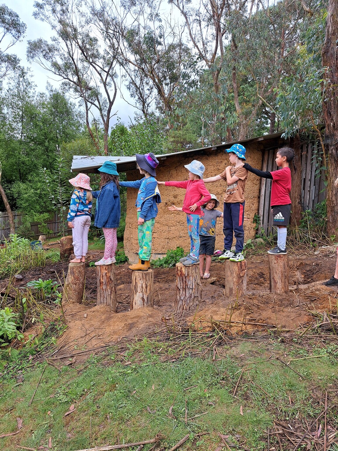 Kindlehill children balance together on stumps, supporting one another and connecting to Country within their bushland environment.