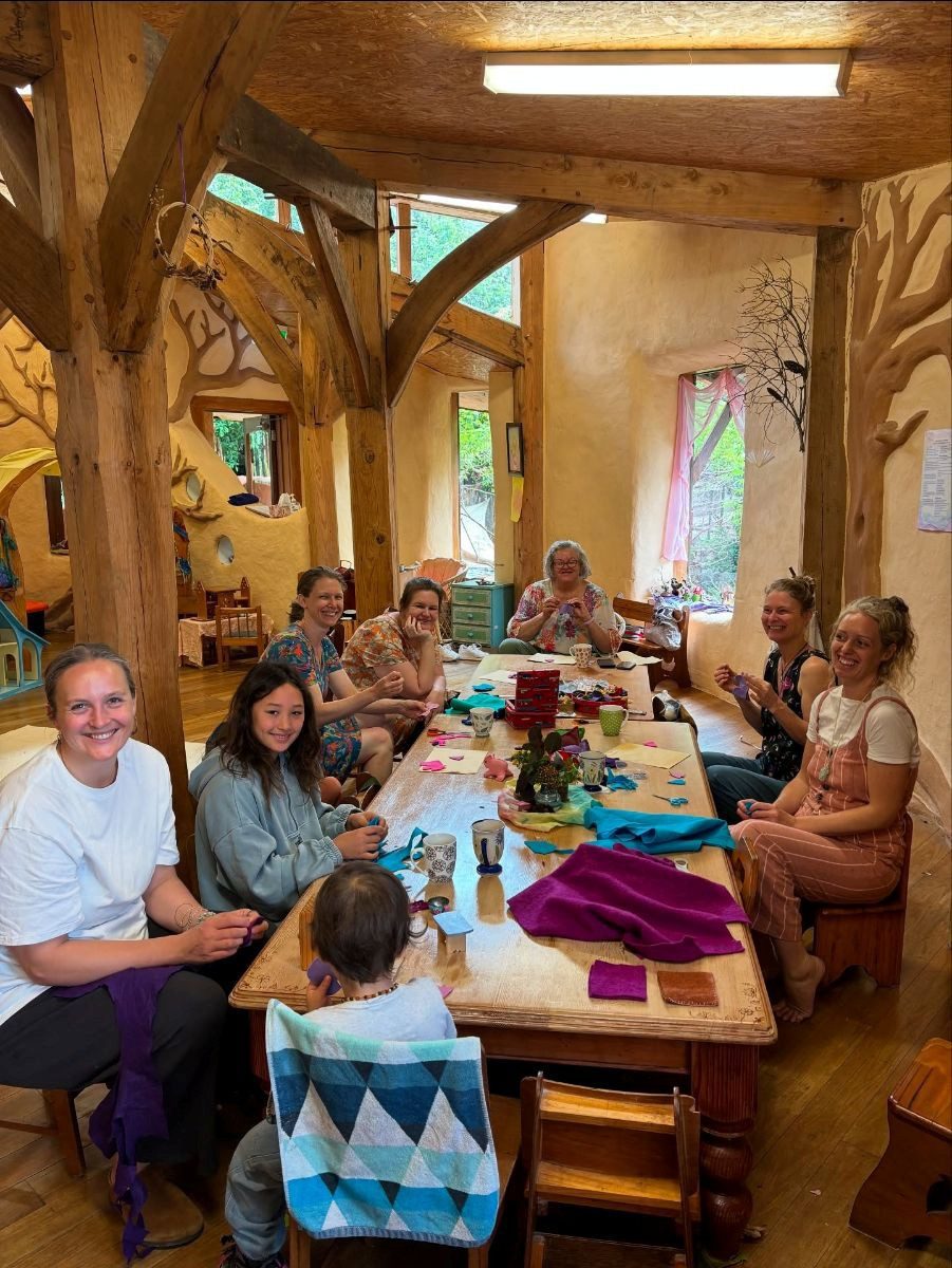 Community members share stories and craft around a rustic table, fostering connection within the warm Kindlehill Kindy hearth.