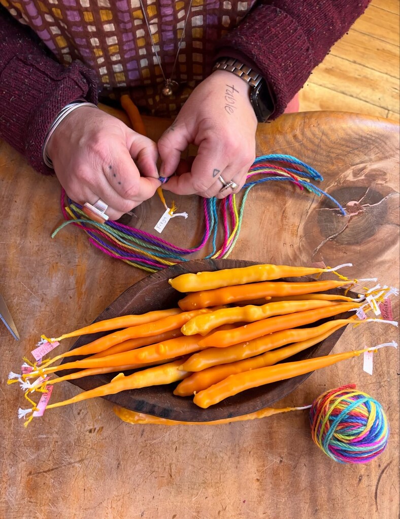 Artistic hands tie rainbow yarn to beeswax candles, honouring the community through rhythmic craft and heart-led connection.